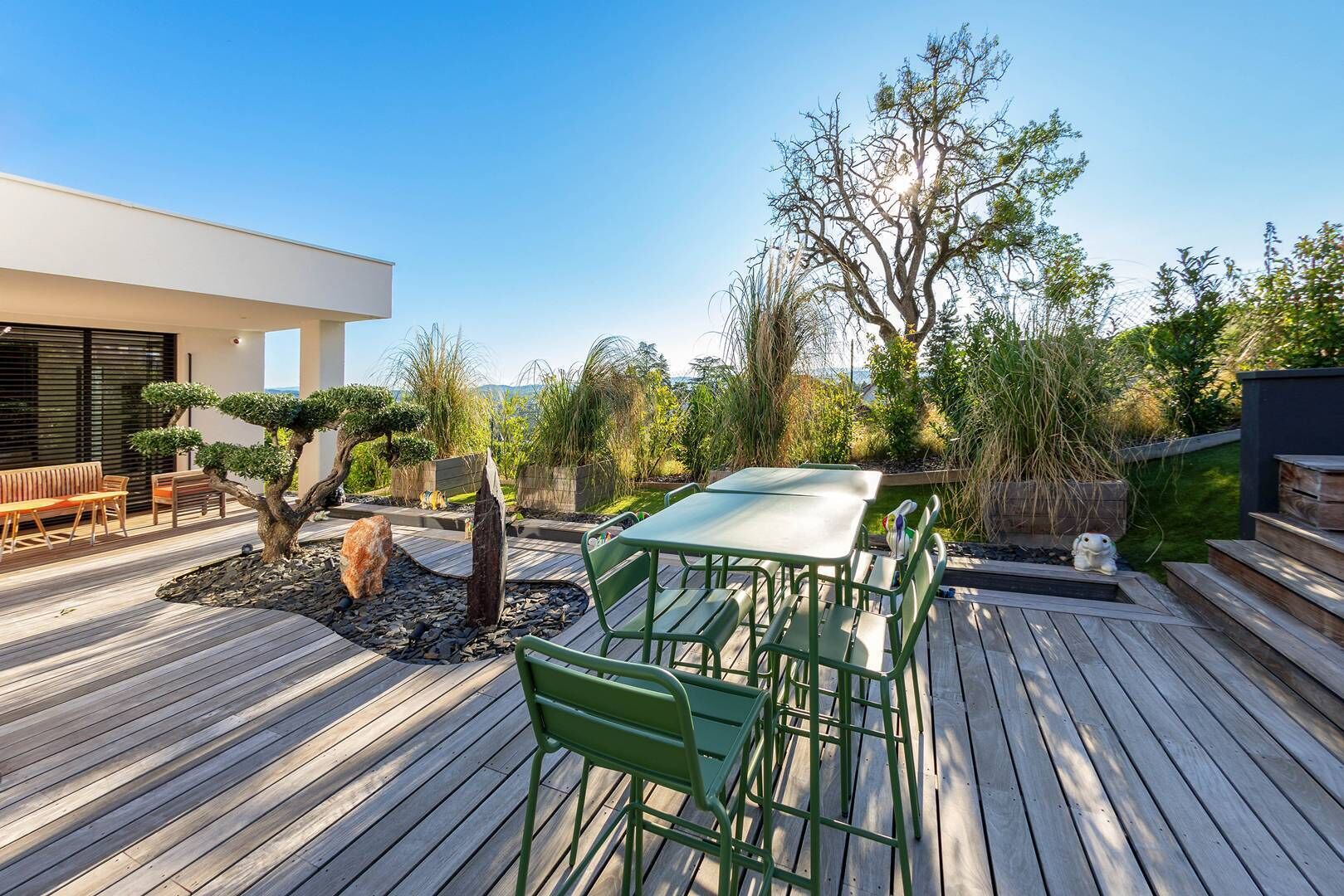 A wooden deck with a table and chairs, surrounded by a garden with various plants and a bonsai tree.