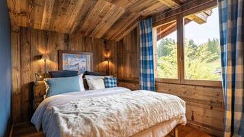 Un dormitorio acogedor con techo de madera, una cama grande con muchas almohadas, una mesita de noche con lámpara y una ventana con cortinas.