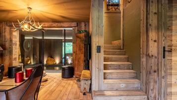 Un intérieur rustique avec un escalier en bois, un coin repas agrémenté d’un lustre, et une vue sur l’extérieur à travers une porte vitrée.