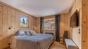 A wooden-paneled bedroom with a bed, a bench, a TV, and a window with a view of a building and trees.