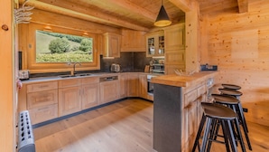 A wooden cabin-style kitchen with a large window, wooden cabinets, a central island with stools, and a hanging light fixture.
