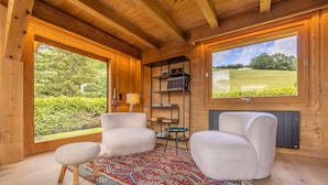A cozy room with wooden walls, large windows, and a view of a green landscape. There are two white chairs, a small stool, and a patterned rug.