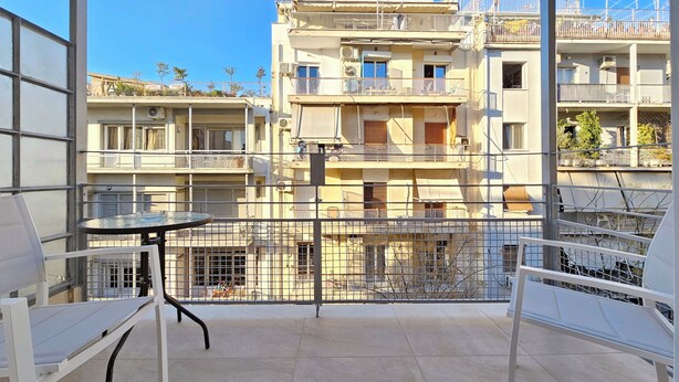 En balkon med bord og stole, med udsigt over en høj beboelsesejendom med både balkoner og gelændere.