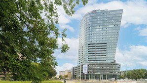 Een modern hoog gebouw met een gebogen gevel, waarschijnlijk een hotel of kantoor, met een heldere lucht en wat groen op de voorgrond.