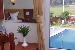 A hotel room with a bed, a bedside table with a plant, a mirror, and a view of a swimming pool. - Hotel Yatra Sauraha (Sauraha)