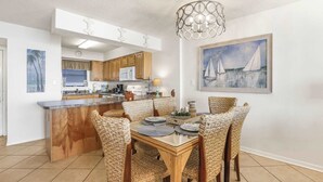 A modern kitchen with wooden cabinets, a dining area with wicker chairs, and a painting of sailboats on the wall. - Seaside Beach 5417 | Beachfront Condo w/ Pool, Sauna & Balcony Gulf (Orange Beach)