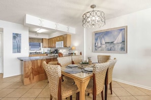 A modern kitchen with wooden cabinets, a dining area with wicker chairs, and a painting of sailboats on the wall. - Seaside Beach 5417 | Beachfront Condo w/ Pool, Sauna & Balcony Gulf (Orange Beach)