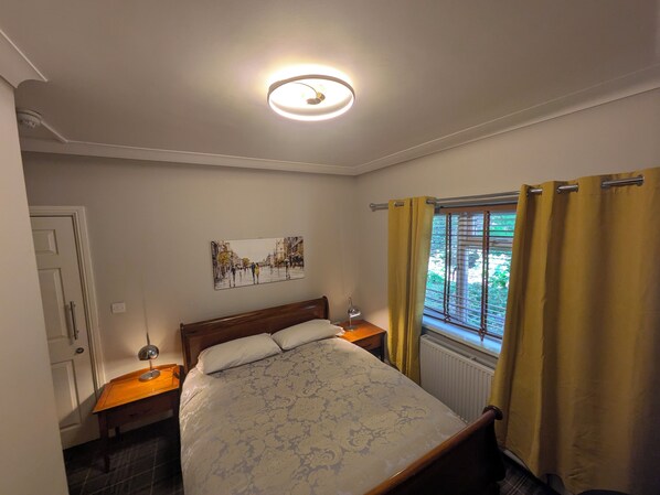 A bedroom with a bed, a bedside table, a lamp, a window with curtains, and a wall-mounted picture. - The Godalming Hotel (Godalming)