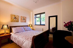A bedroom with a bed, bedside lamps, a window with blinds, a mirror, and a vase with flowers.