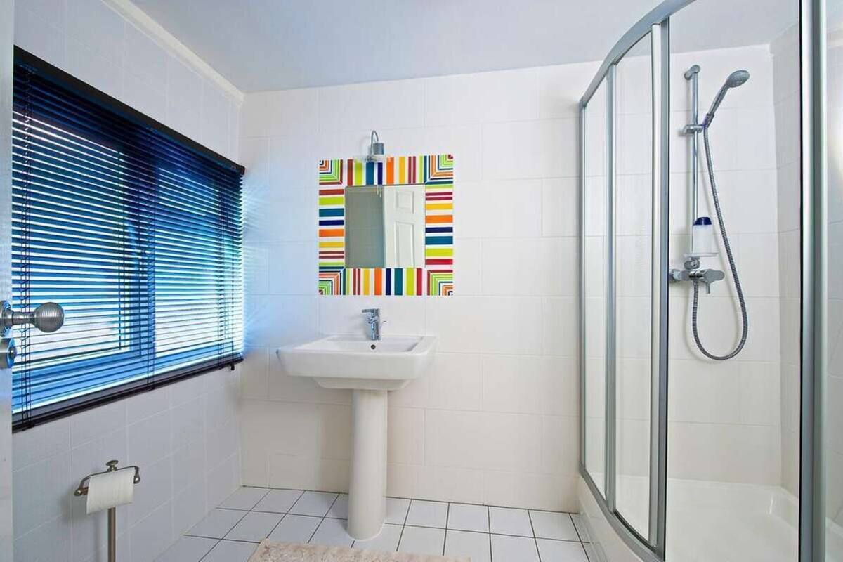 Une salle de bain avec un lavabo, un miroir doté d’un cadre coloré, une douche et une fenêtre avec des stores.