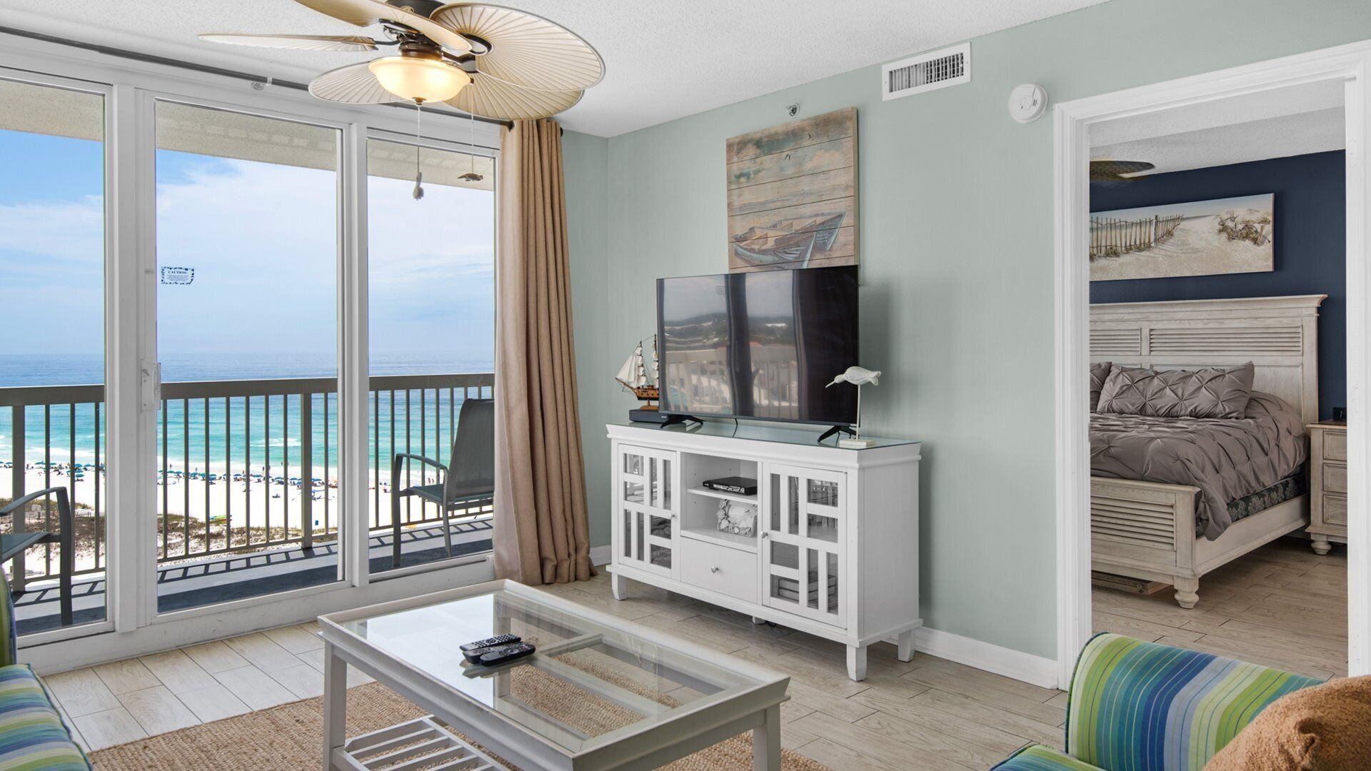 Ein Zimmer mit Balkon und Blick auf einen Strand, ein Bett mit Kopfteil, ein Fernseher auf einer Kommode und ein Couchtisch.