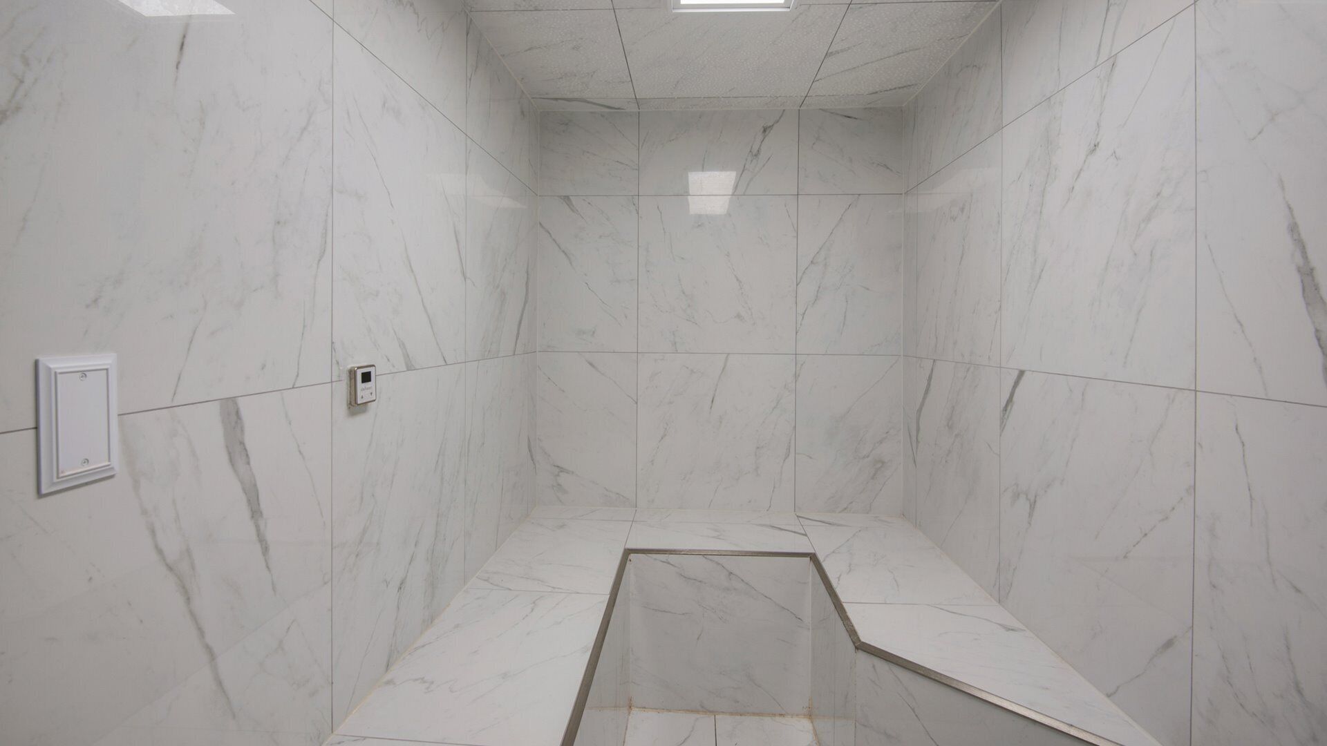 A bathroom with white marble walls and floor, a shower area with a drain, and a light switch on the wall.
