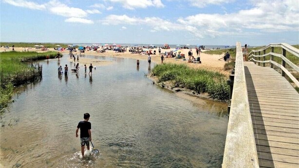 En strandscene med en træpromenade der fører ned til vandet, folk der bader, og strandgæster i baggrunden.