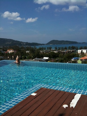 A swimming pool with clear blue water, surrounded by a wooden deck. In the background, there are buildings and hills.
