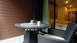 A small round table with a white mug and a plate of croissants, accompanied by wicker chairs, positioned next to a sliding glass door leading to a bedroom with a bed and a window with blinds. - Luxurious 45sqm Apartment in Peaceful Patong Location (Patong)