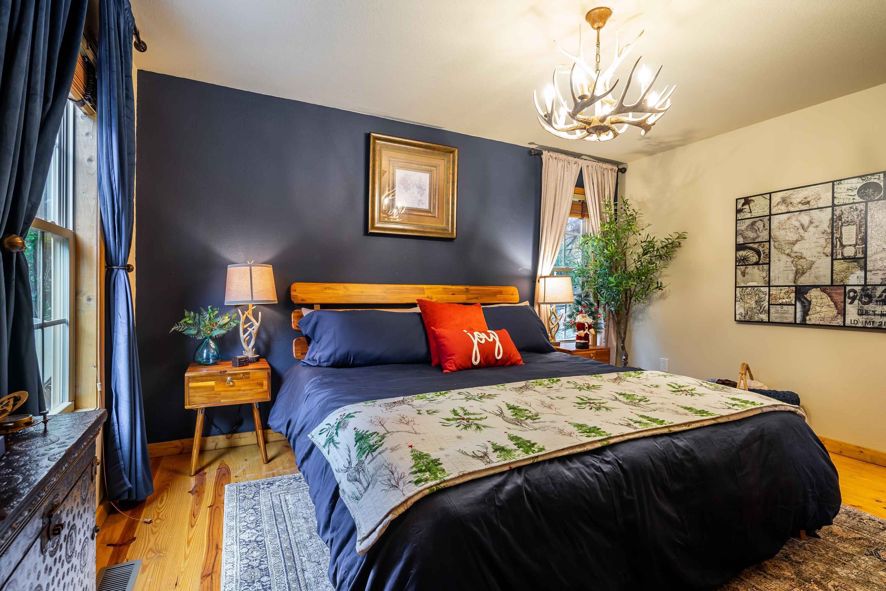 A bedroom with a dark blue bed, a wooden headboard, a red pillow, a framed picture above the bed, a chandelier, a plant, and a world map on the wall.