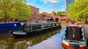 A narrow canal with two narrowboats docked, a brick building, and a bridge in the background.