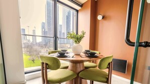 A dining area with a round table, four chairs, a vase with greenery, and a city view through large windows.