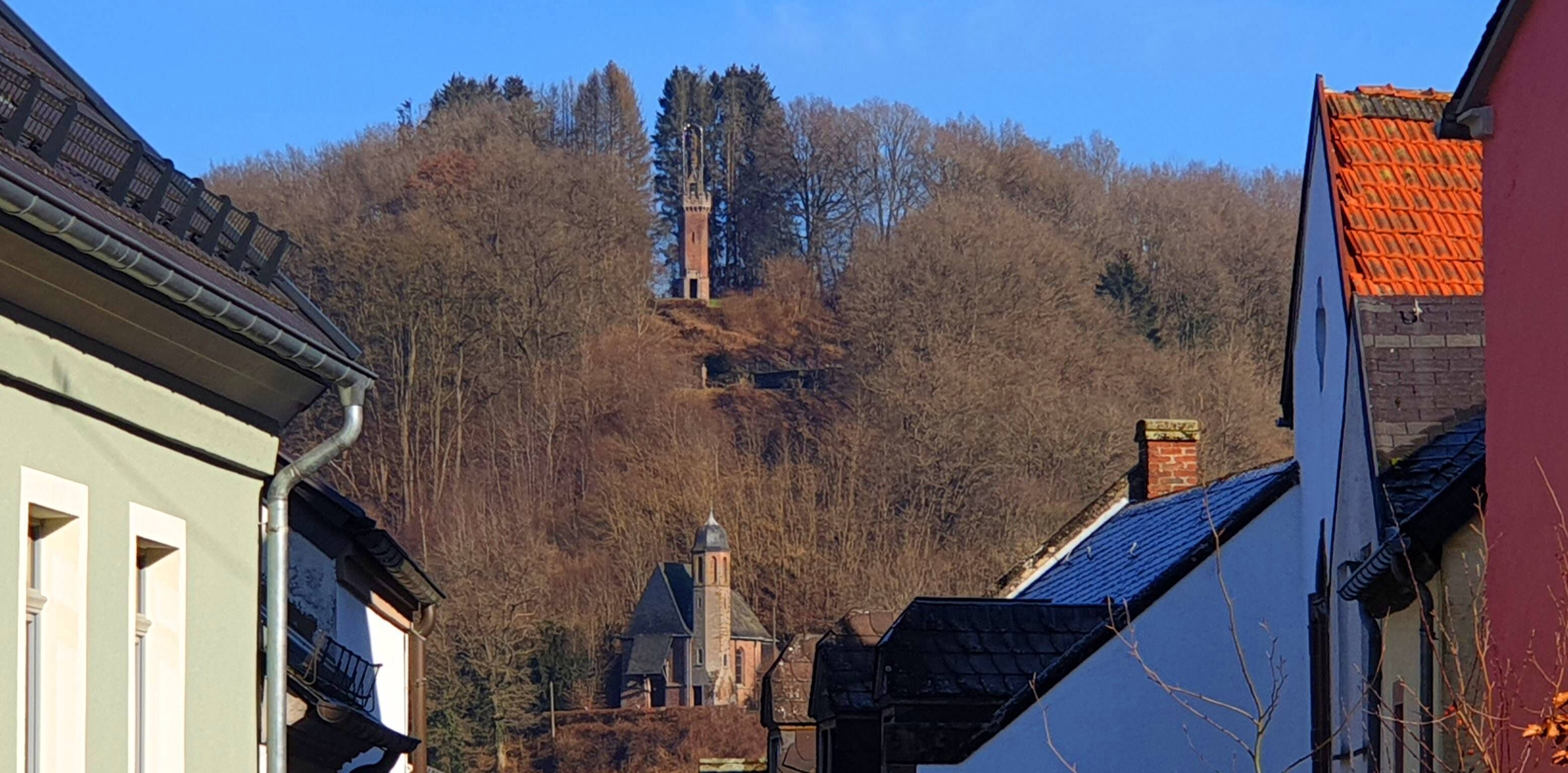 A building with a steep roof and a chimney, surrounded by trees and other buildings.