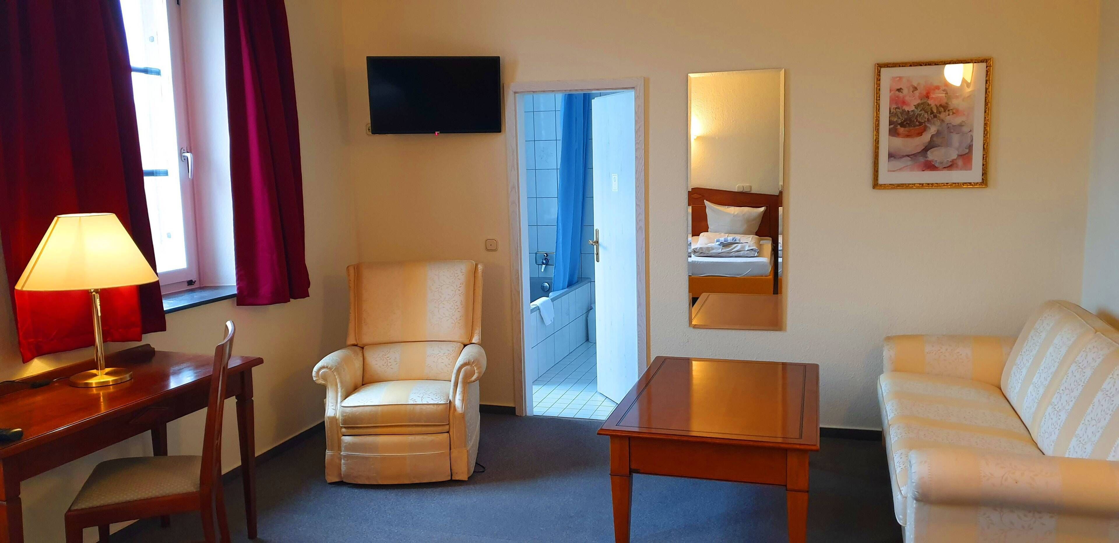 Habitación de hotel con sofá, una silla, una mesa, televisión y puerta que da a un balcón.
