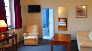 Habitación de hotel con sofá, una silla, una mesa, televisión y puerta que da a un balcón.