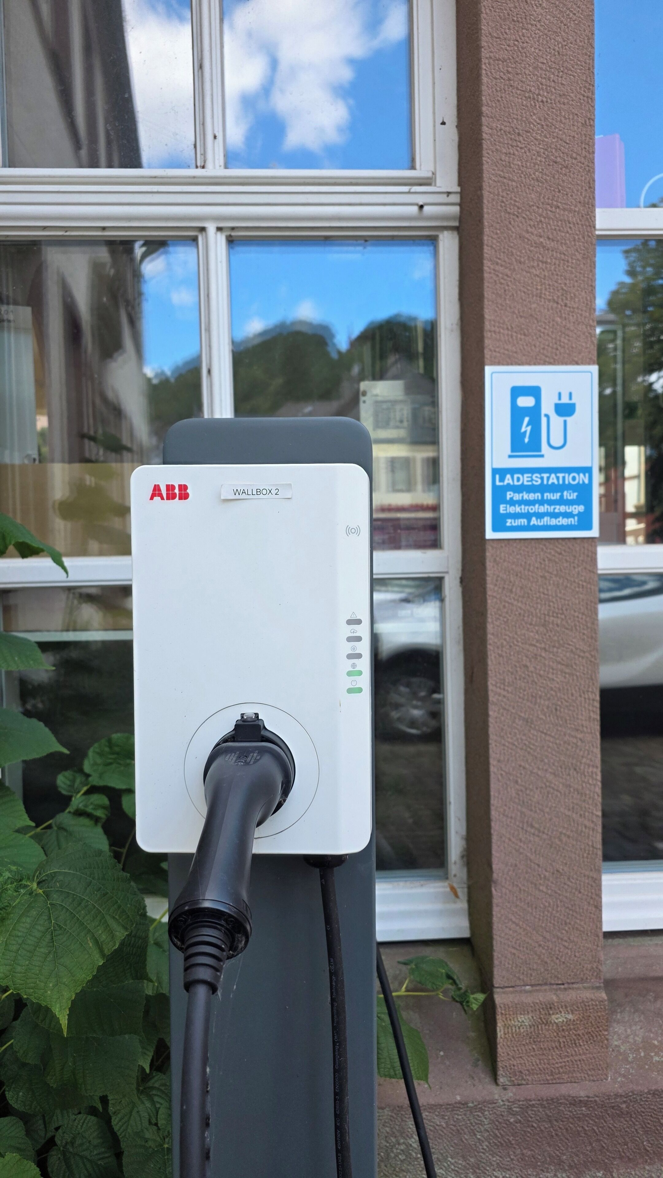 An electric vehicle charging station labeled 'ABB WALLBOX 2' with a plug inserted, a sign indicating 'LADESTATION' for electric vehicles, and a window reflecting a car and trees.