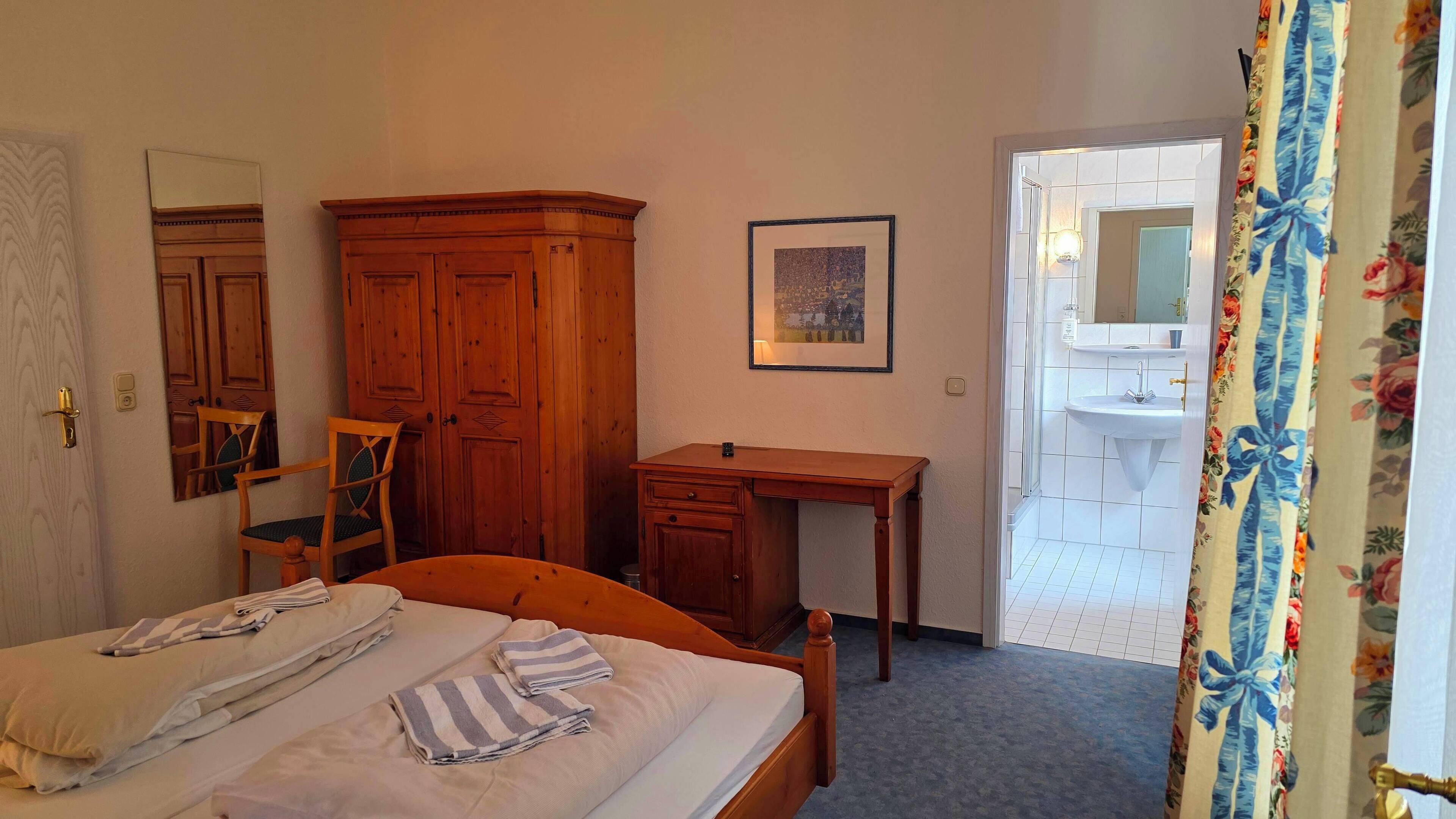 A bedroom with a bed, a wooden desk, a chair, a wardrobe, a framed picture, and a bathroom visible through an open door.
