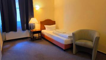 Hotellihuone, jossa on sänky, yöpöytä lampun kanssa, tuoli ja ikkuna, jossa on verhot.