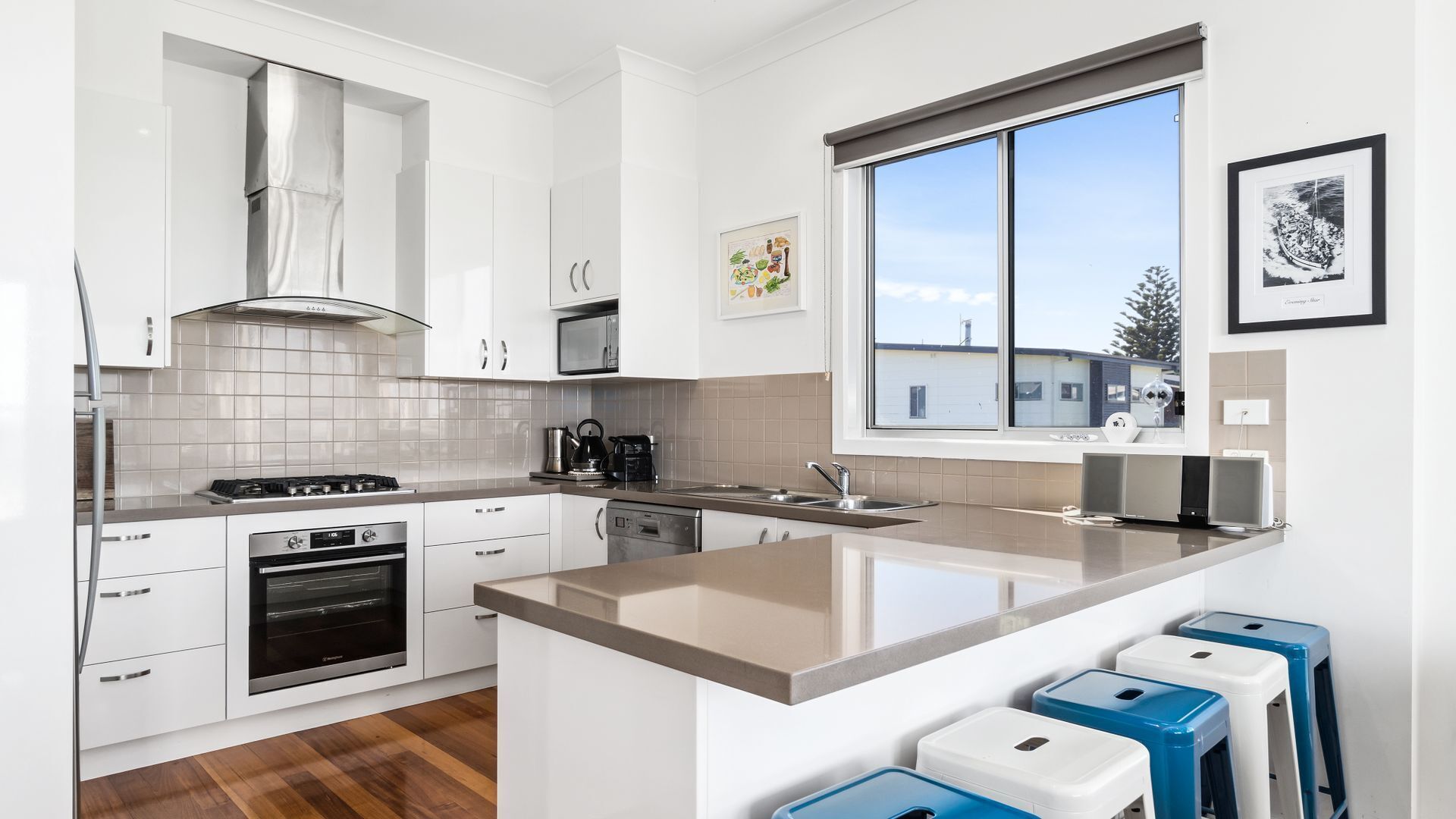 Una cocina moderna con armarios blancos, una campana extractora de acero inoxidable, horno empotrado, cafetera y ventana con vista a edificios.