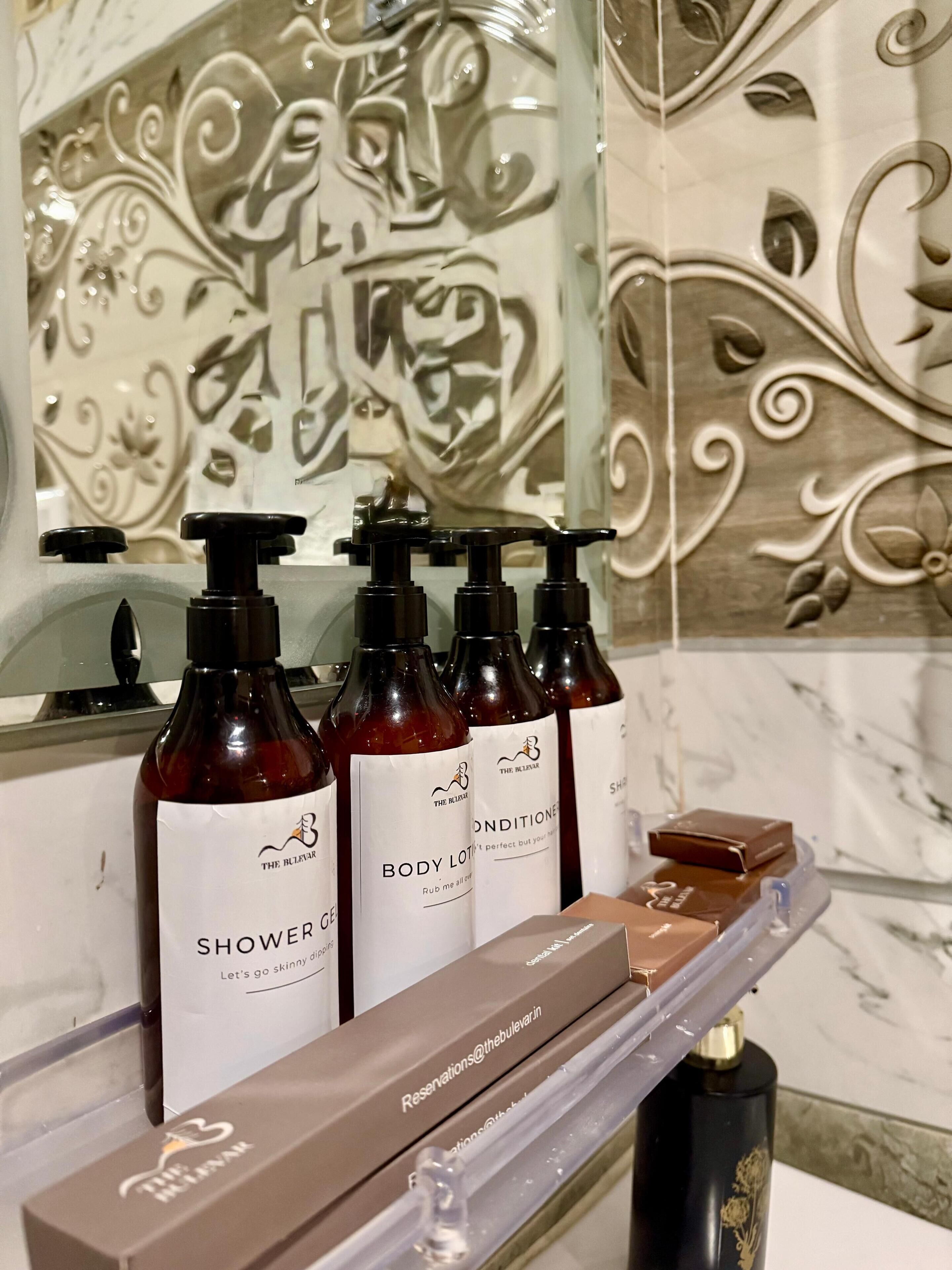 A bathroom with a marble wall and a mirror. There are bottles of shower gel and body lotion on a shelf.