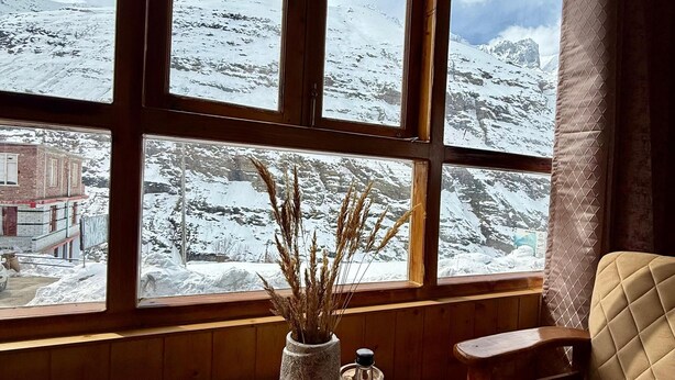 A room with large windows offering a view of a snowy mountain landscape. - The Bulevar-Kunsil (Lahul)