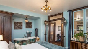 A bedroom with a large bed, a freestanding bathtub, a vanity with a mirror, and a chandelier.