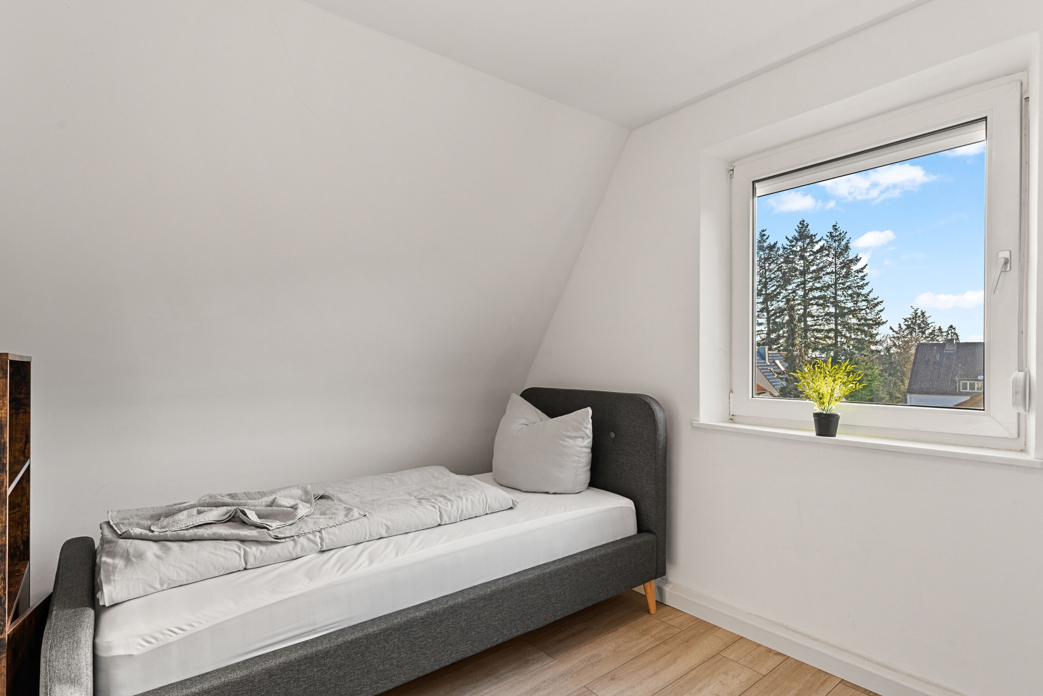 A small, single-bed room with a sloped ceiling and a window overlooking a tree.
