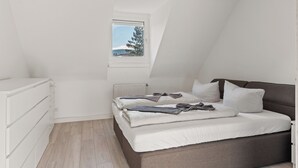 Een slaapkamer met een schuine kap, een raam met uitzicht op bomen, een bed met wit beddengoed en een witte dressoir.