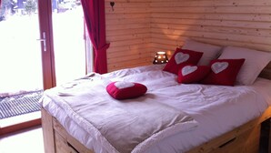 Un dormitorio de cabaña de madera con una cama, almohadillas rojas con forma de corazón y una ventana con cortinas.