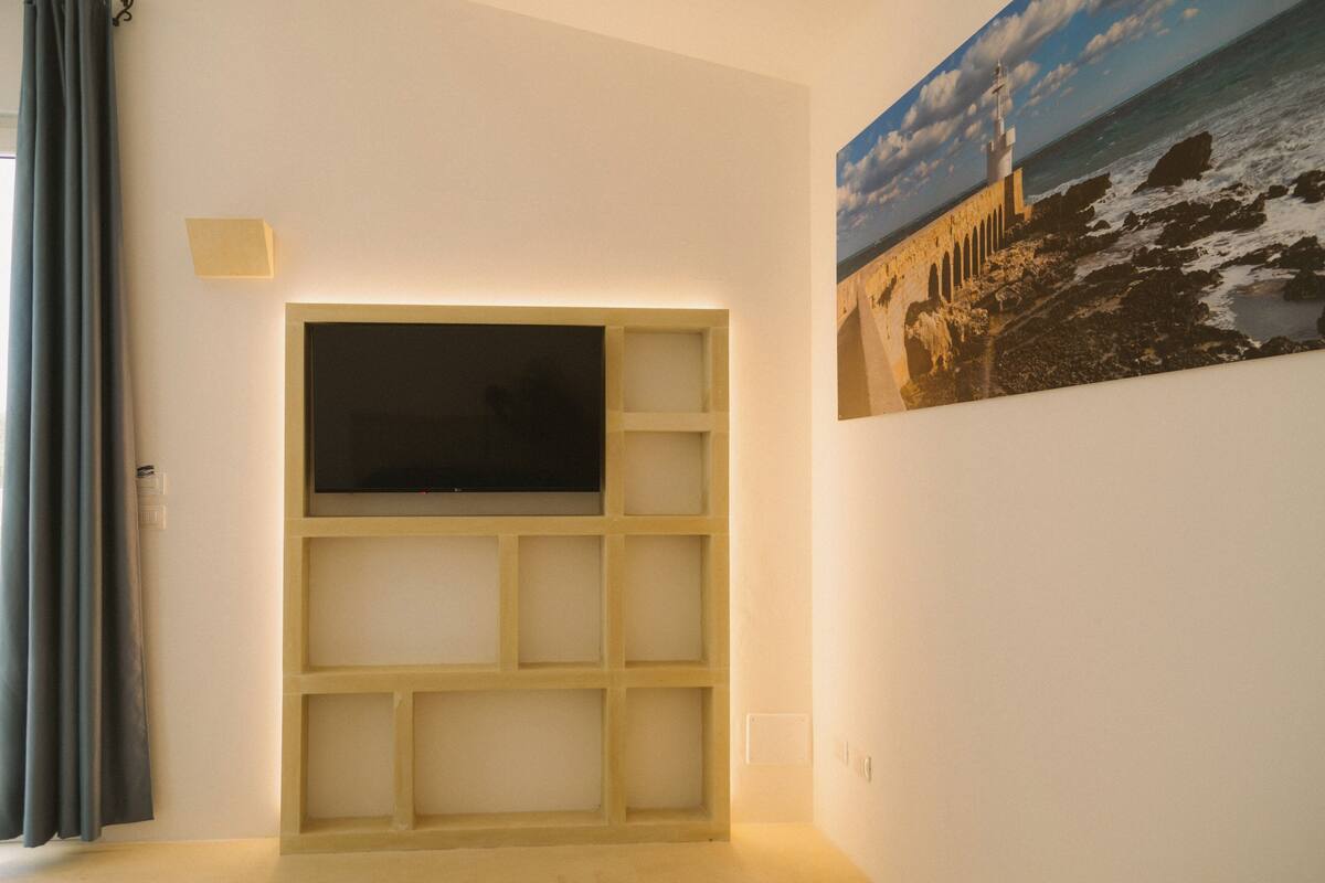 Una habitación con una televisión montada en un estante de madera, un cuadro de un faro en la pared y una cortina en la ventana.