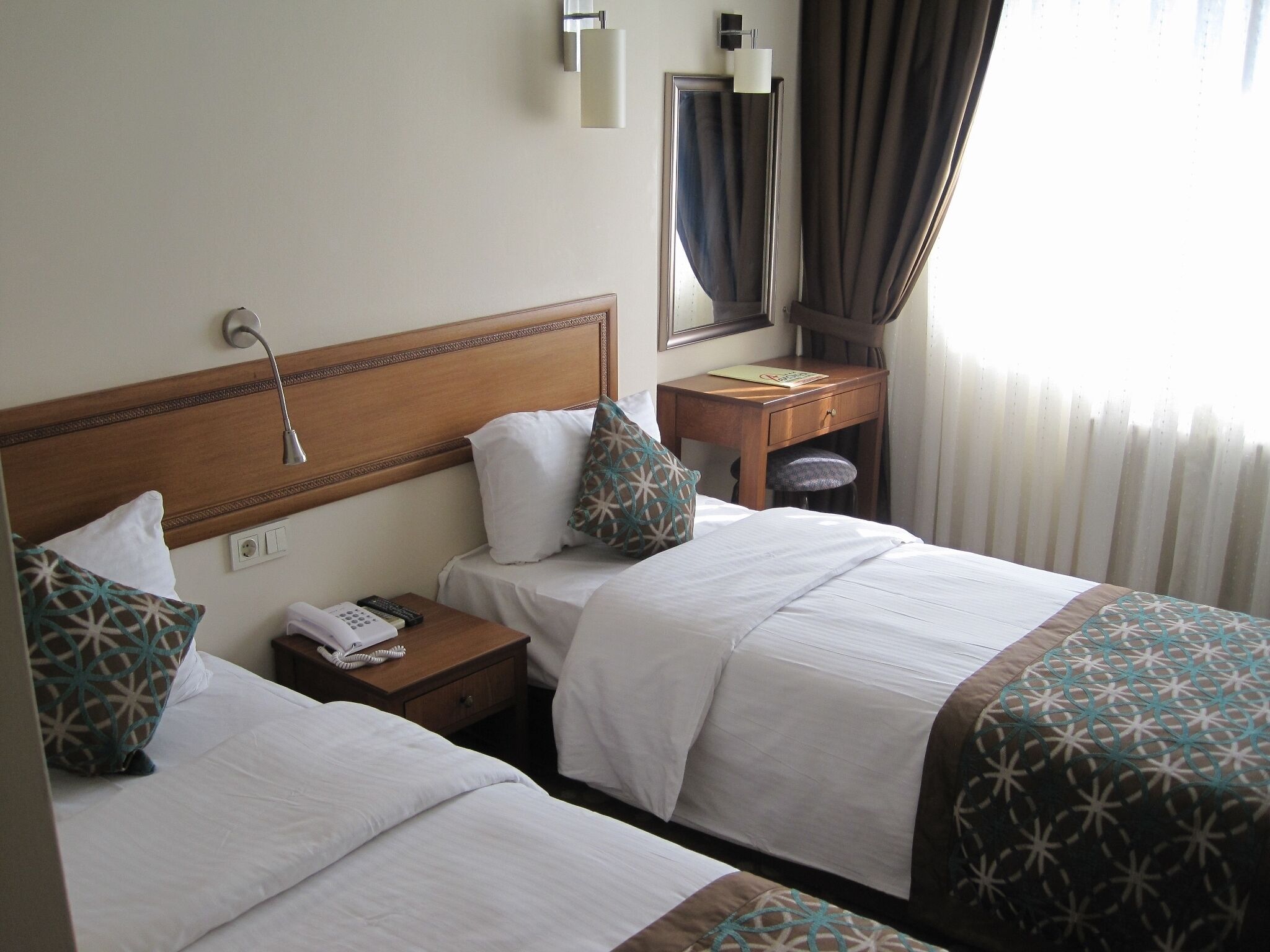 A hotel room with two beds, a nightstand with a telephone and remote controls, a small wooden desk, and a mirror on the wall.