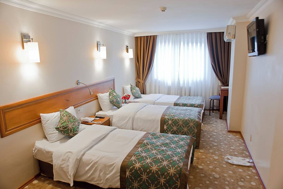 A hotel room with two beds, a wooden headboard, a small table, a chair, a TV, and a window with curtains.