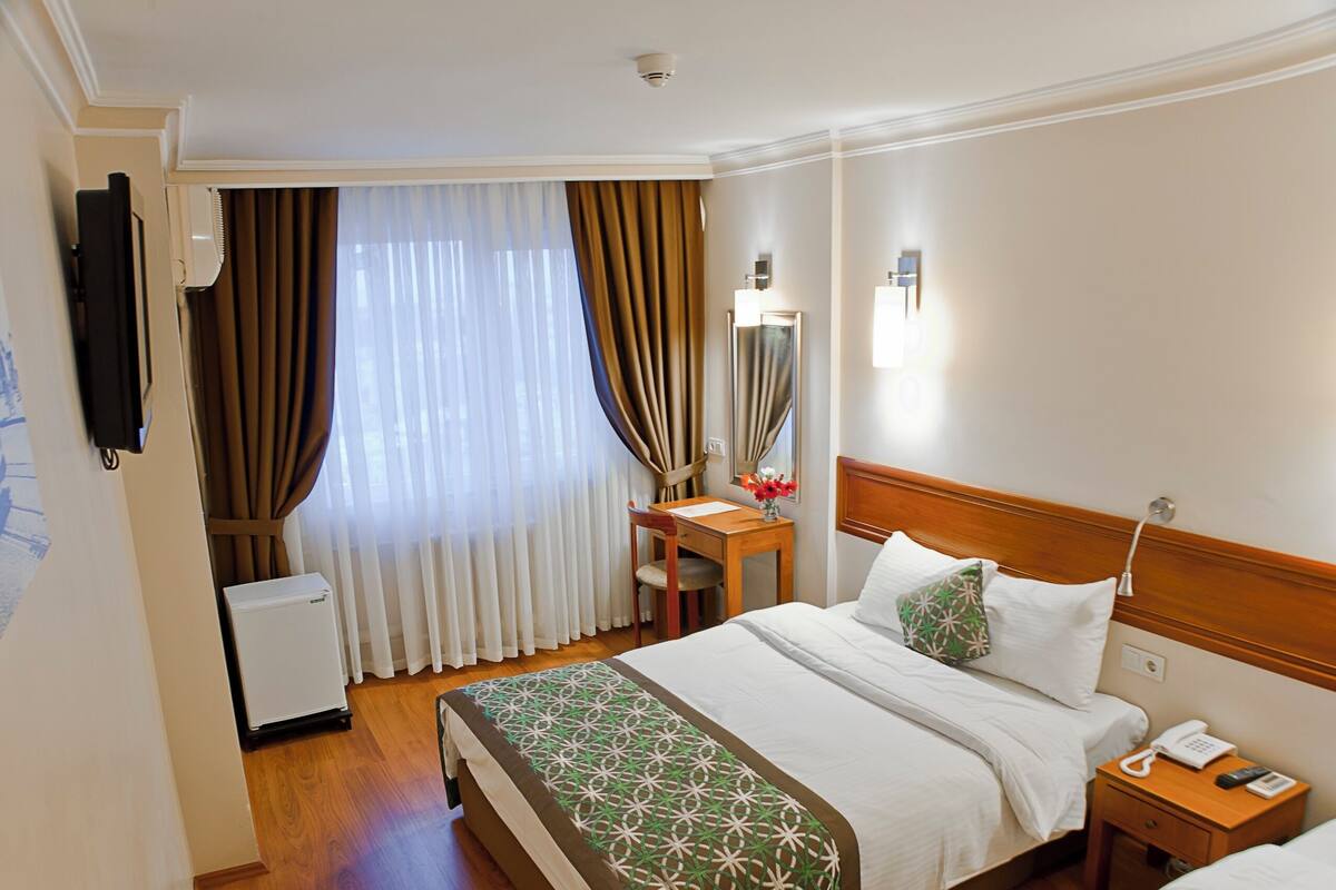A hotel room with a bed, bedside table, lamp, and a small desk with a chair.