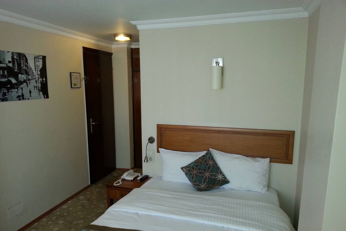 A hotel room with a bed, a bedside telephone, a wall-mounted light, and a door leading to a hallway.