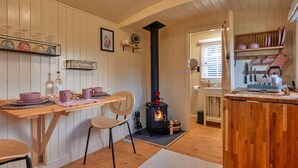 A cozy kitchen with wooden walls, a central wood-burning stove, a dining table with chairs, and shelves with kitchenware.