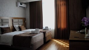 Habitación de hotel con dos camas, cabecero de madera, unidad de aire acondicionado, mesita de noche con teléfono y un jarrón de flores, y ventana con cortinas.