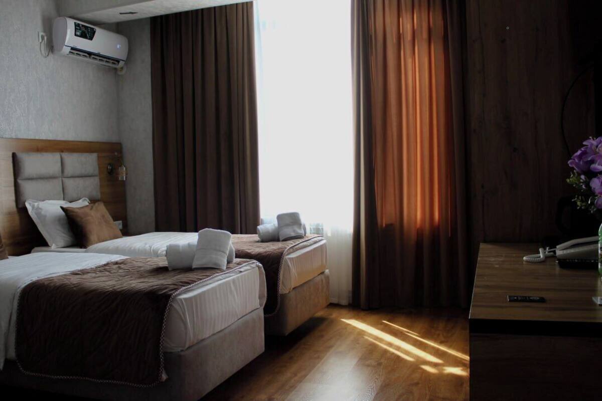 A hotel room with two beds, a wooden headboard, an air conditioning unit, a bedside table with a phone and a vase of flowers, and a window with curtains.