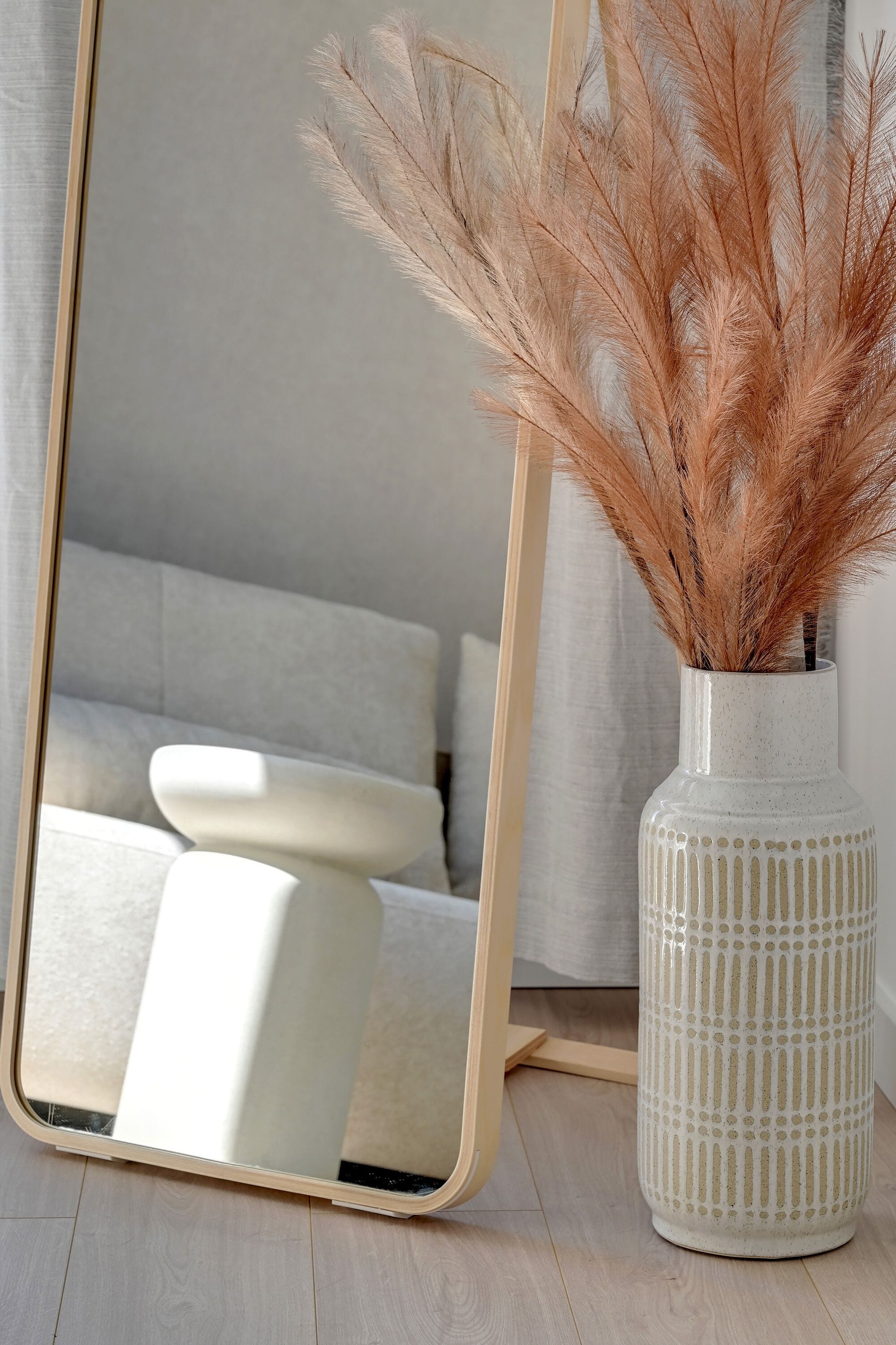 Un miroir avec un cadre beige, un vase blanc contenant des plumes d’herbe des pampas et une chaise blanche.