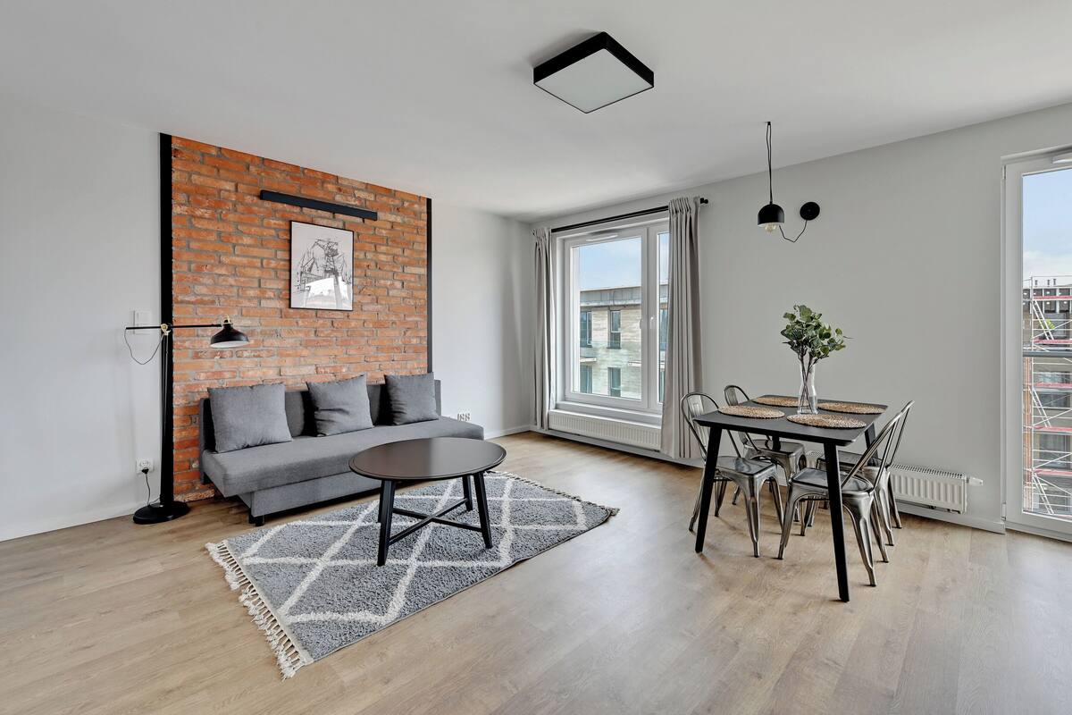 Ein modernes Wohnzimmer mit einem grauen Sofa, einem runden Couchtisch und einem Esstisch mit Stühlen. Eine Wand mit Backsteinoptik, ein gerahmtes Bild, ein großes Fenster mit Vorhängen und eine Heizung sind ebenfalls zu sehen.