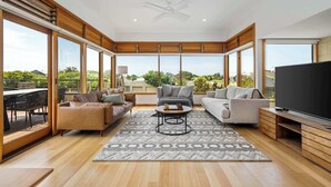 A spacious living room with a large TV, comfortable seating, a ceiling fan, and large windows offering a view of the outdoors. - Ocean Breeze - Retreat in Torquay, Sleeps 11! (Torquay)