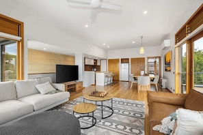 A modern living room with a sofa, coffee table, and TV. There is a kitchen area in the background with a dining table and chairs. - Ocean Breeze - Retreat in Torquay, Sleeps 11! (Torquay)