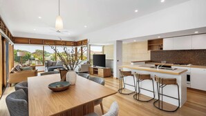A modern kitchen with a central island, a dining area with a wooden table, and a living space with sofas and a TV. - Ocean Breeze - Retreat in Torquay, Sleeps 11! (Torquay)