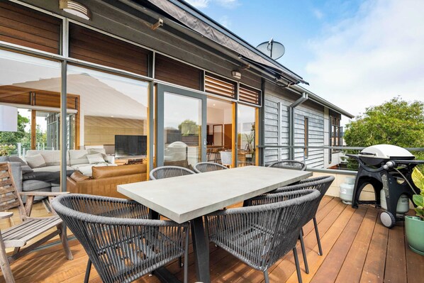Une terrasse extérieure moderne, dotée d’une table et de chaises pour les repas, d’un barbecue et d’une grande porte-fenêtre coulissante donnant sur un espace de vie intérieur.