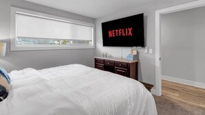 Ein Schlafzimmer mit einem großen Bett, einem an der Wand montierten Fernseher, einem Fenster mit Jalousien, einem Schminktisch und einer Tür.